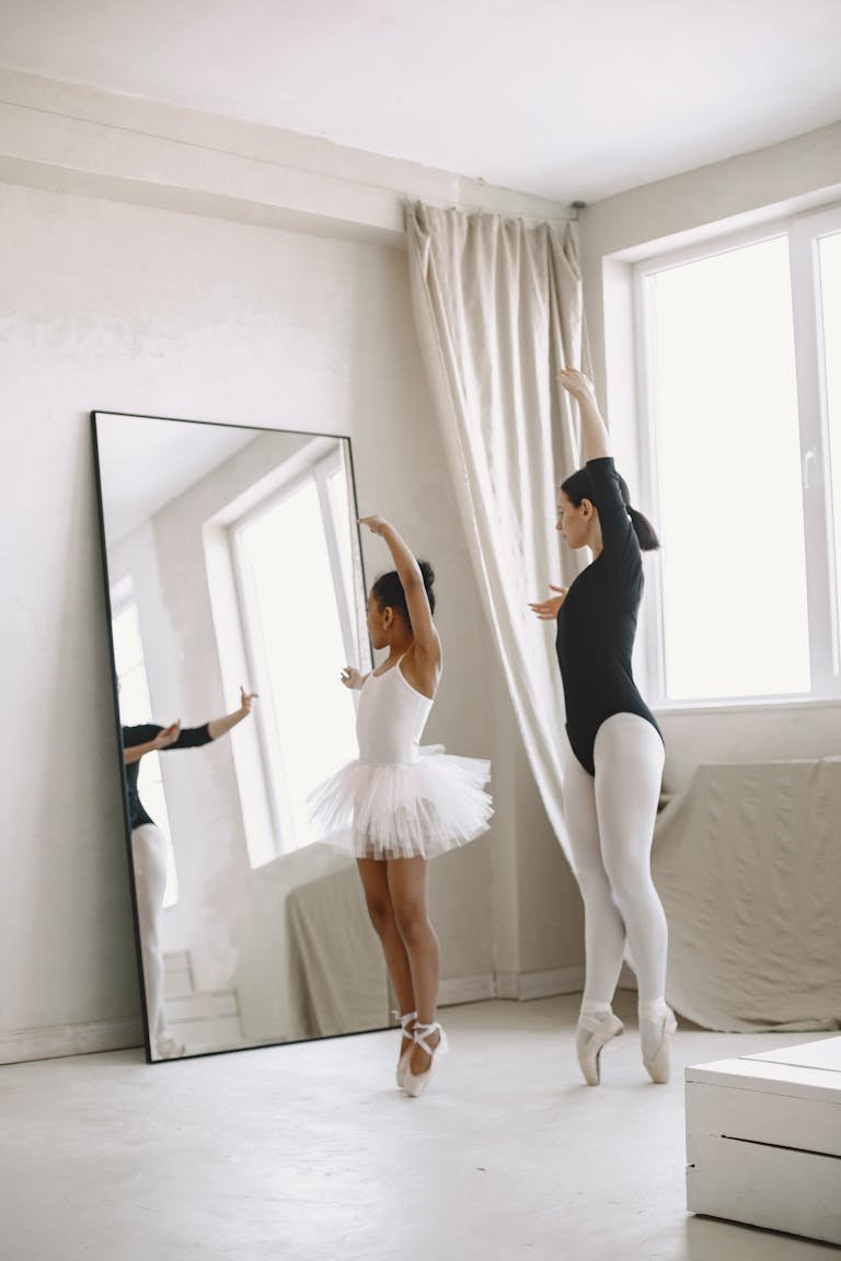 A young girl practices ballet with a teacher in a bright dance studio.